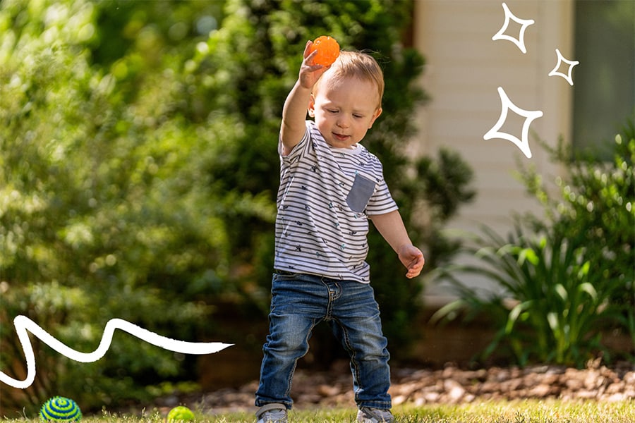 18-month-old toddler practices throwing a ball outside.