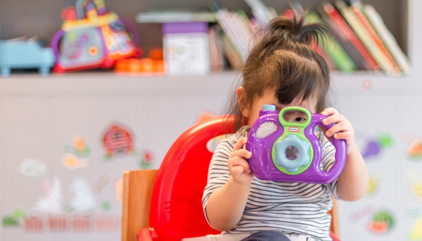 Toddler girl plays with toy camera at early childhood education center.