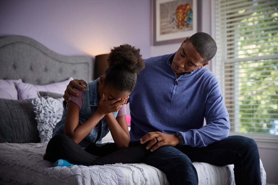 Preteen cries on her bed while dad comforts her by putting his arm around her.