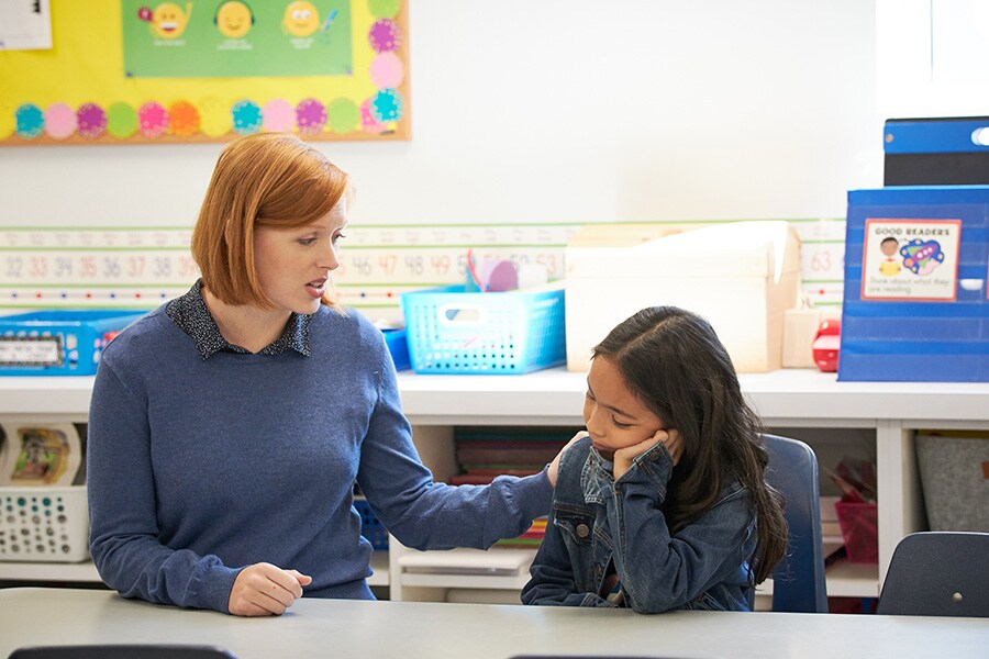 A photo of a woman teacher comforting an elementary school-aged girl slumped at her desk