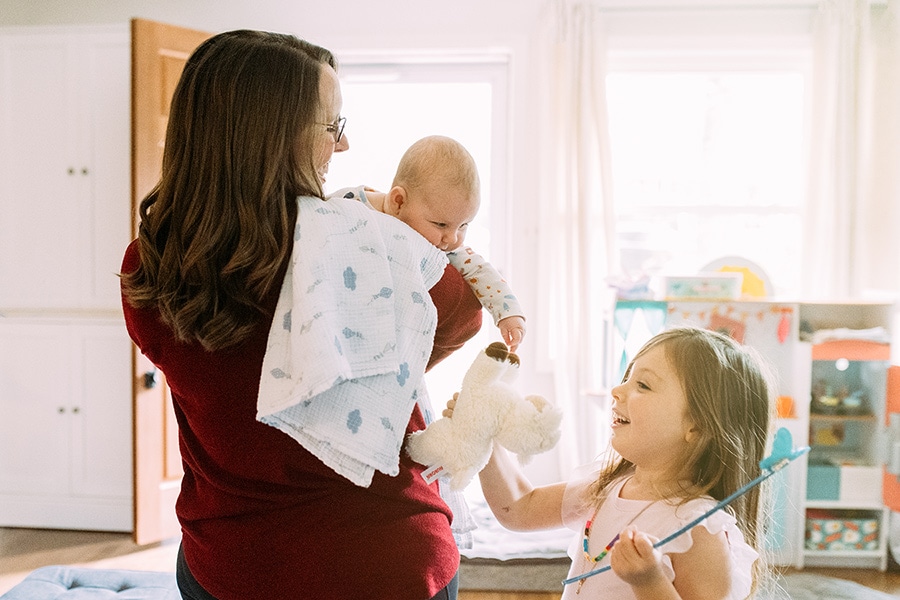 Mom holding baby with older sibling offering baby a stuffed animal toy, smiling