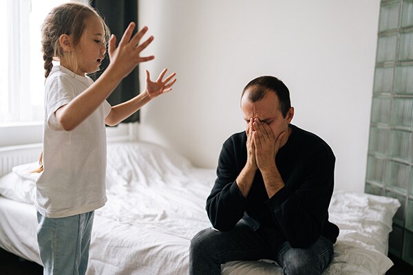 A dad sits on his school-aged daughter's bed feeling overwhelmed while she yells at him.