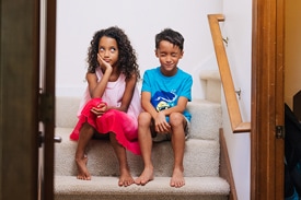 A school-age brother and sister look frustrated with each other while sitting on stairs at home