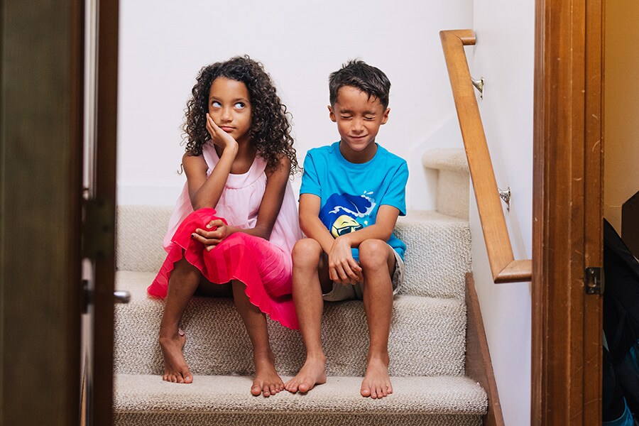 A school-age brother and sister look frustrated with each other while sitting on stairs at home