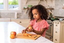Toddler peels orange by herself at kitchen countertop.