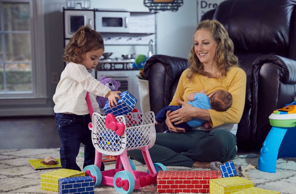 Mother breastfeeds newborn while toddler girl plays