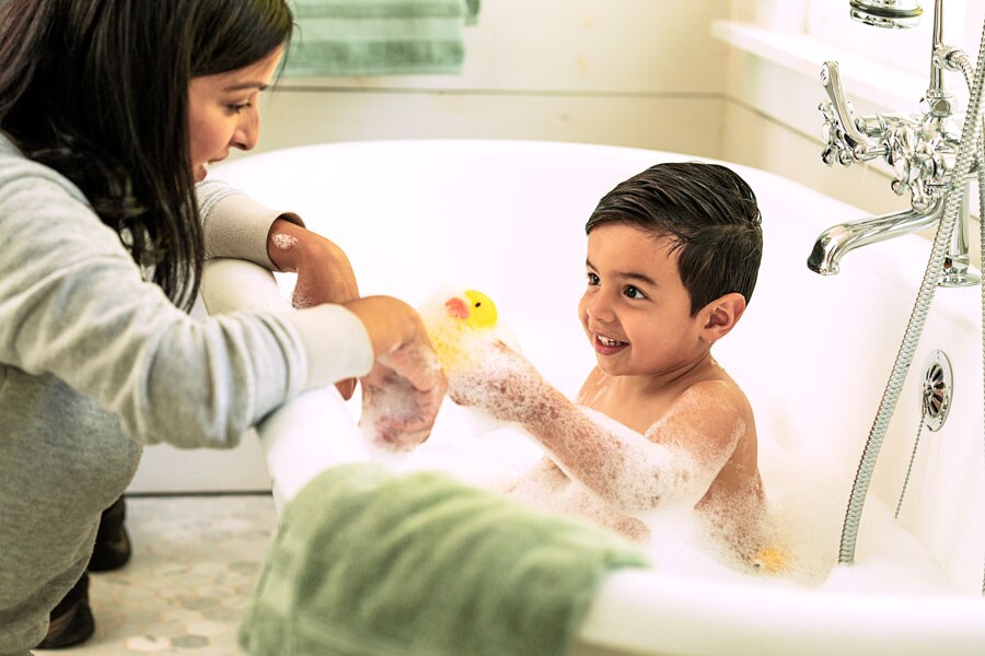 Caregiver tests water temperature for safety while preparing a bath with toys and bubbles.