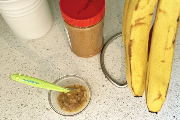 A photo of a banana and peanut butter mixture on a countertop with a baby spoon, a jar of peanut butter and bananas.