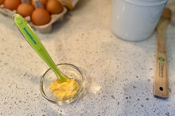 A photo of a hard-boiled egg mashed and mixed with infant formula on a counter with a baby spoon and carton of eggs.