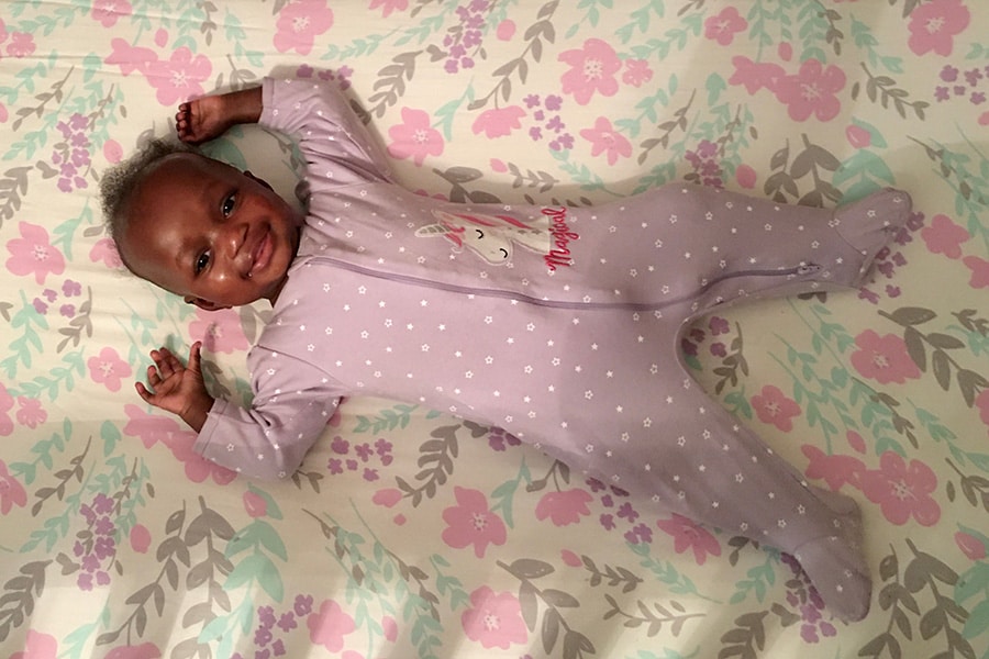An infant lays alone, on her back, in a clear crib while wearing a purple onesie
