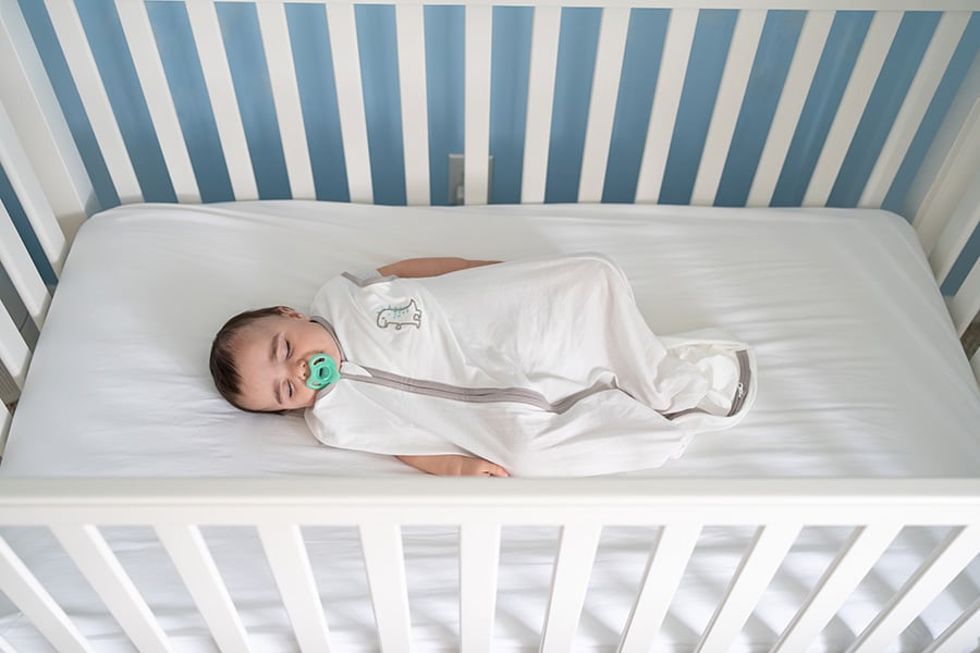 A photo of a baby boy sleeping soundly on his back in a sleep sack in an empty crib. 