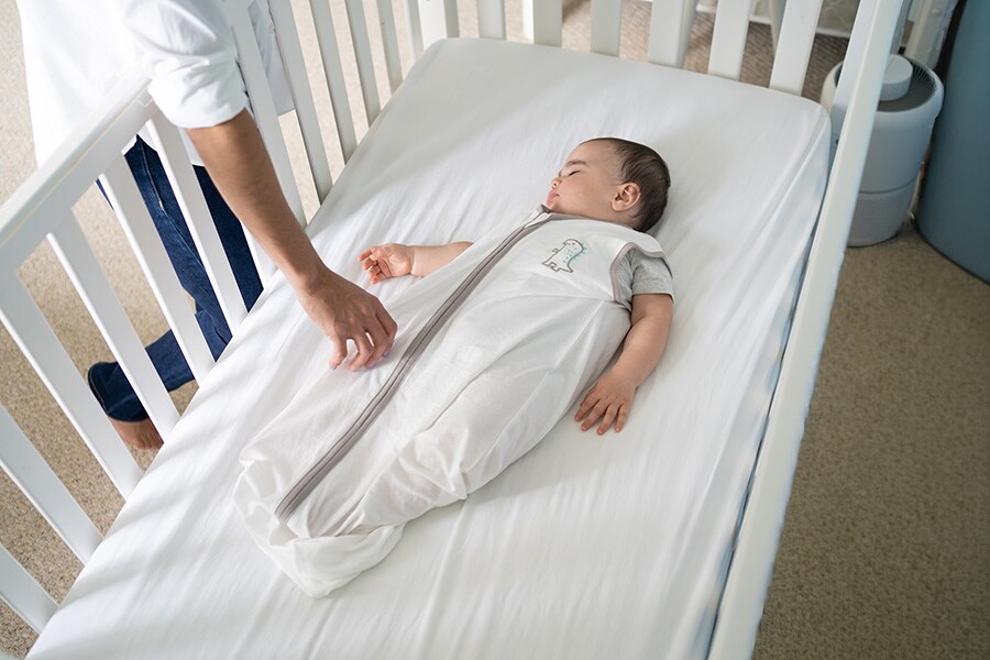 A photo of a parent reaching down into a crib where their baby is sleeping soundly in a sleep sack. 