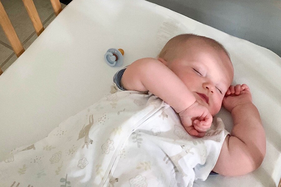 Infant sleeping alone, on their back, in a clear crib with a pacifier nearby. 