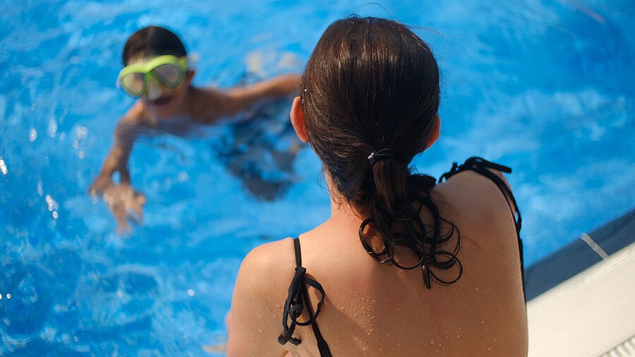A mother sitting on side of pool, acting as a water watcher while her son swims