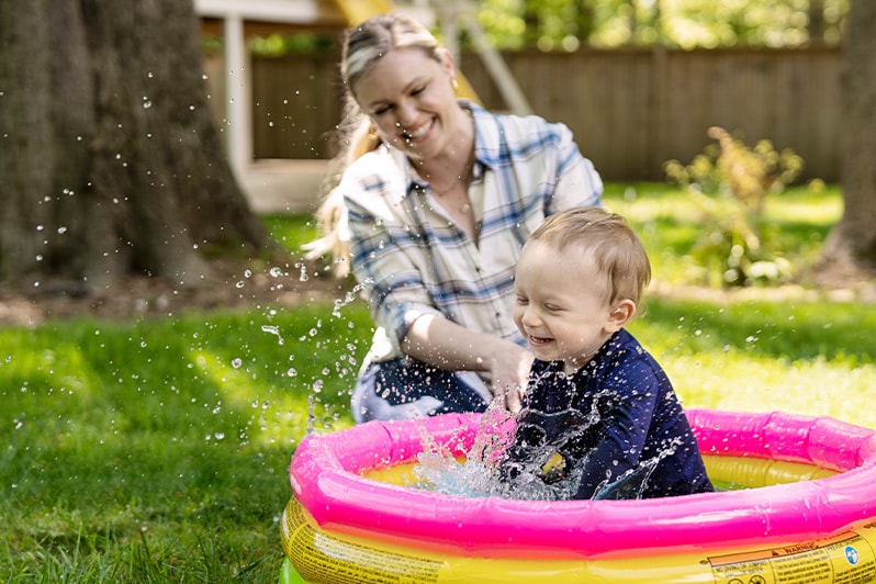 Toddler boy splashes in baby pool while mom uses touch supervision to practice drowning prevention.