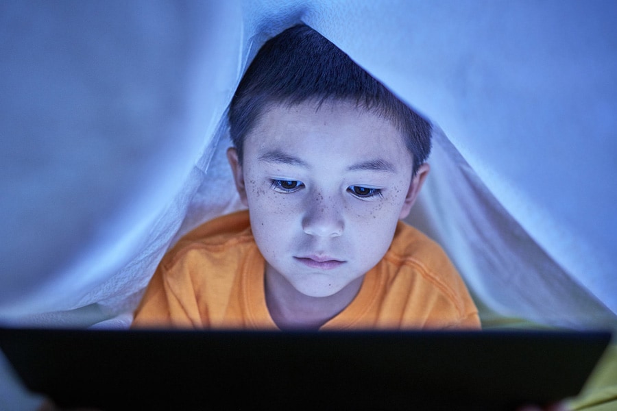 child looking at screen under the covers in bed